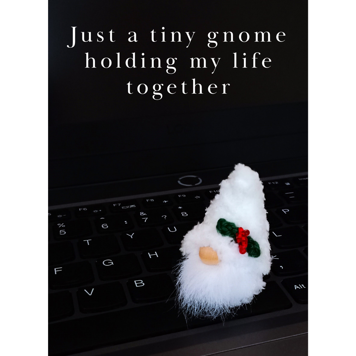 A vertical funny coworker Christmas card featuring an incredibly fluffy white handmade miniature plush gnome sitting on a black laptop keyboard with clean Latin letters. Elegant white text at the top reads: 'Just a tiny gnome holding my life together'.