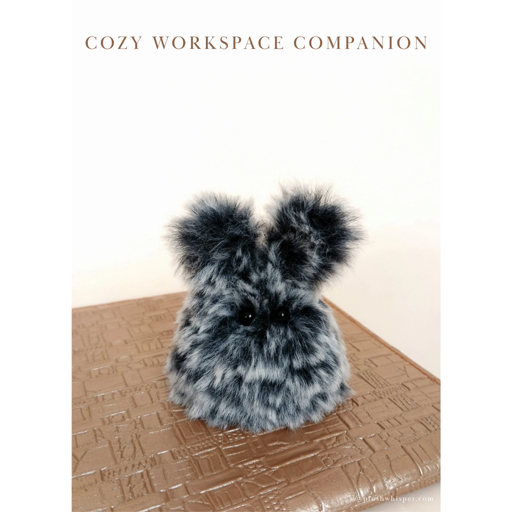 A miniature smoky marbled grey desk bunny handmade from premium tactile faux fur sitting quietly on a gold textured leather notebook. Clean solid light beige studio background with elegant empty space and fine minimalist typography that reads Cozy Workspace Companion at the top. High-end minimalist desk setup aesthetic in quiet luxury wabi-sabi style.
