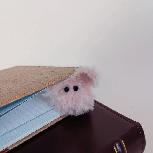 Tiny pink fluffy handmade bunny desk friend peeking out from between a notebook and a dark brown book
