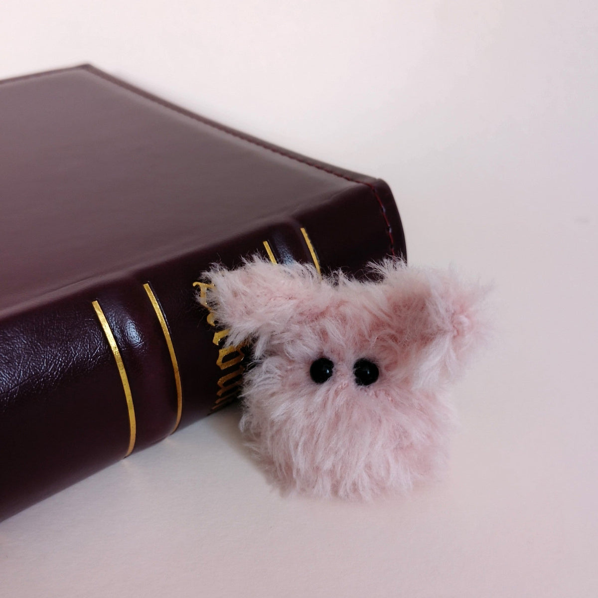 Tiny pink fluffy handmade bunny desk friend sitting beside a dark brown book