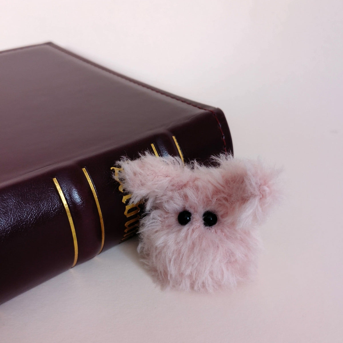 Tiny pink fluffy handmade bunny desk friend sitting beside a dark brown book