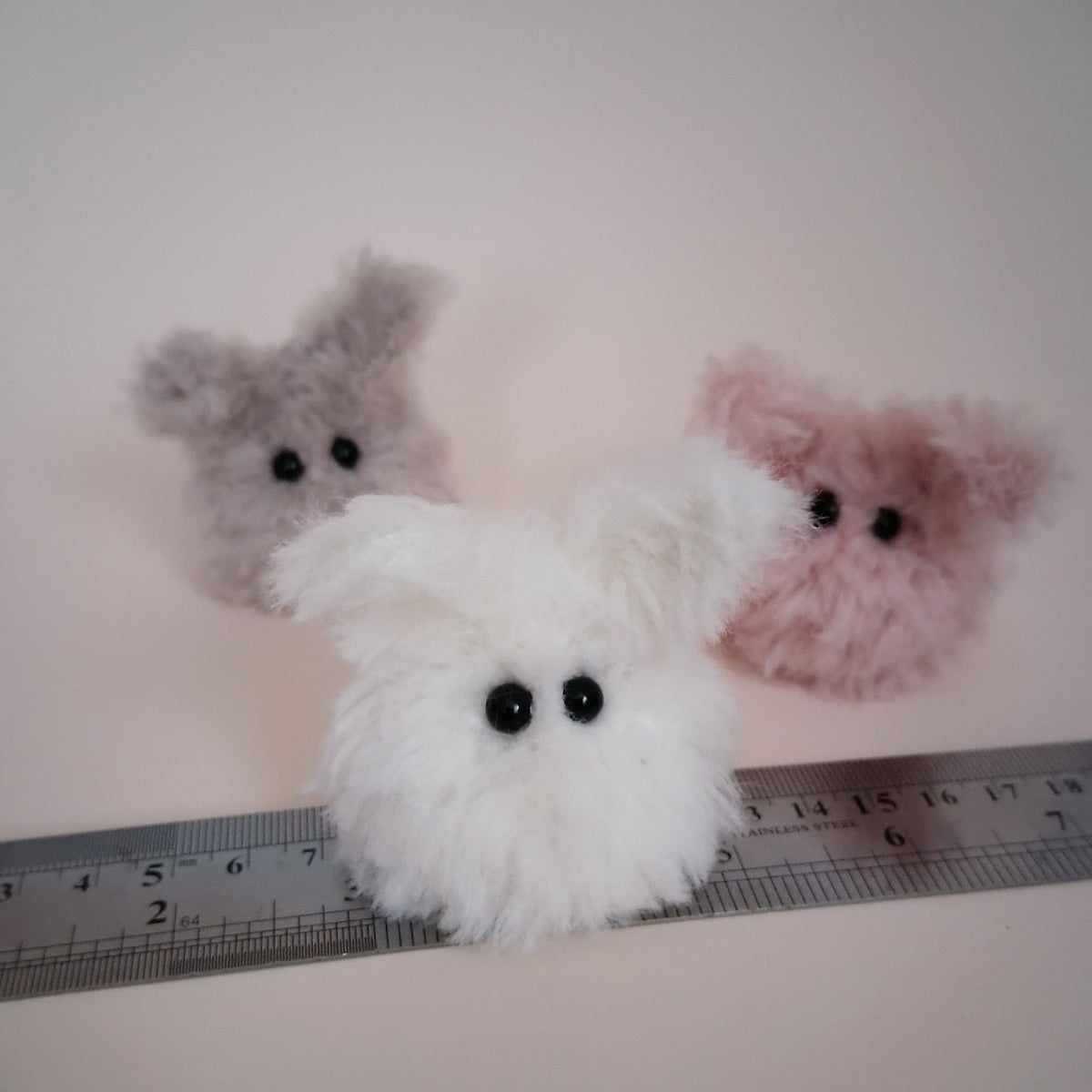 White Worry Bunny handmade from fluffy yarn with gray and pink bunnies behind it, shown beside a ruler for size