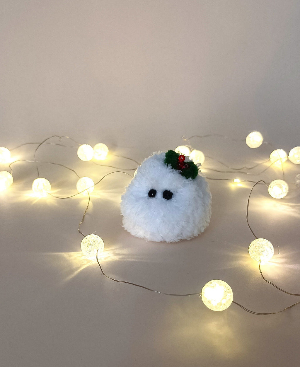 Tiny white crochet Christmas friend with a green holly bow sitting among warm glowing fairy lights on a neutral background.