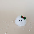 Tiny white crochet Christmas snowball with black eyes and a green holly bow, handmade fluffy desk companion surrounded by pearl decorations.