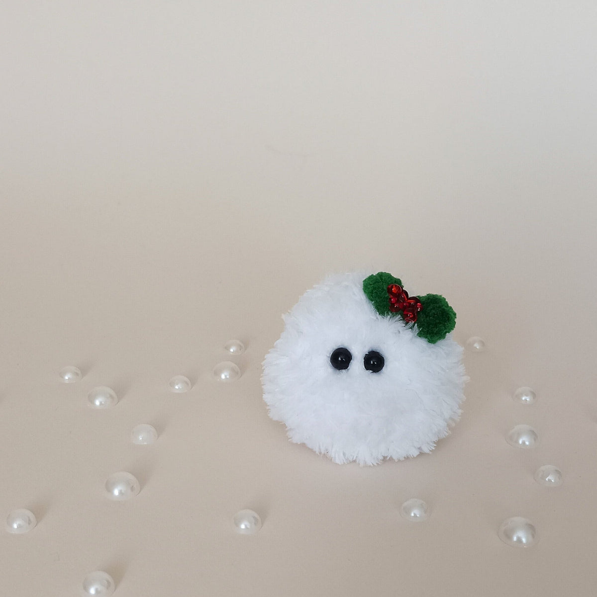 Tiny white crochet Christmas snowball with black eyes and a green holly bow, handmade fluffy desk companion surrounded by pearl decorations.