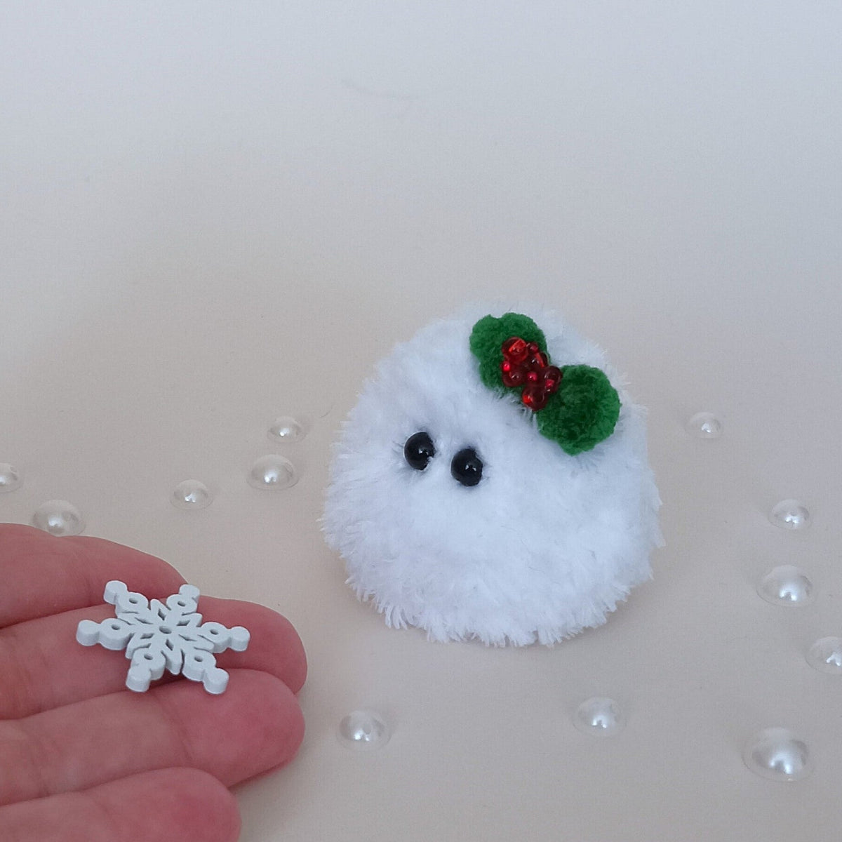 Tiny white crochet Christmas snowball with a green holly bow beside a hand holding a small snowflake, showing the miniature size of the fluffy desk companion.