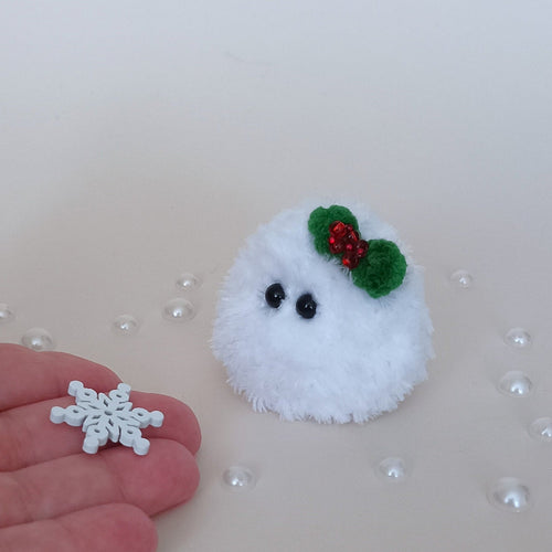 Tiny white crochet Christmas snowball with a green holly bow beside a hand holding a small snowflake, showing the miniature size of the fluffy desk companion.
