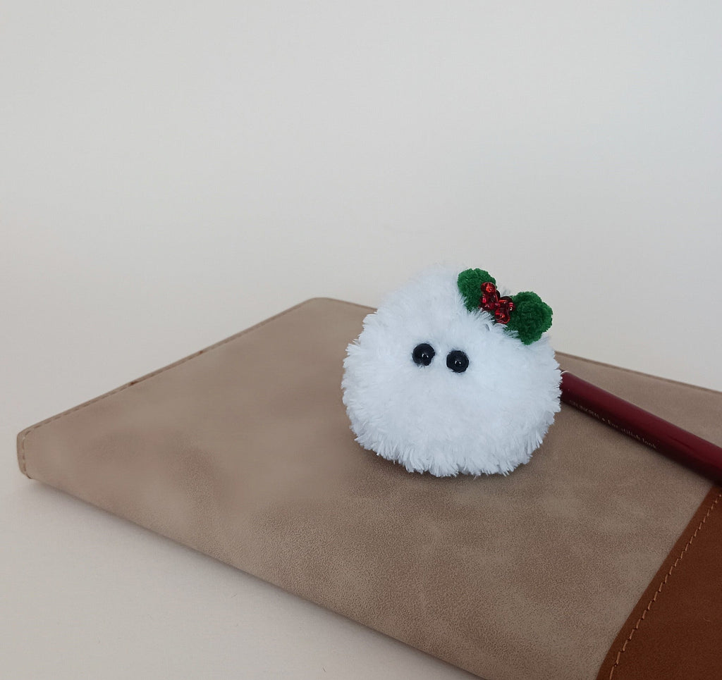 Tiny white crochet Christmas friend with a green holly bow sitting on a neutral notebook beside a red pencil.