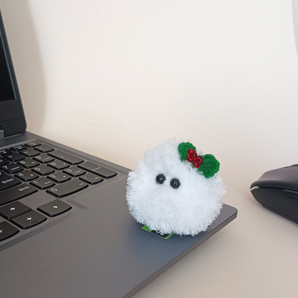 Tiny white crochet Christmas friend with a green holly bow sitting on a laptop beside a computer mouse as a miniature desk companion.