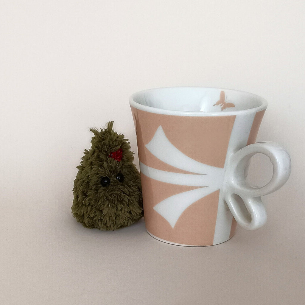 Tiny green crochet Christmas tree with red berry decoration beside a coffee cup, miniature holiday desk companion