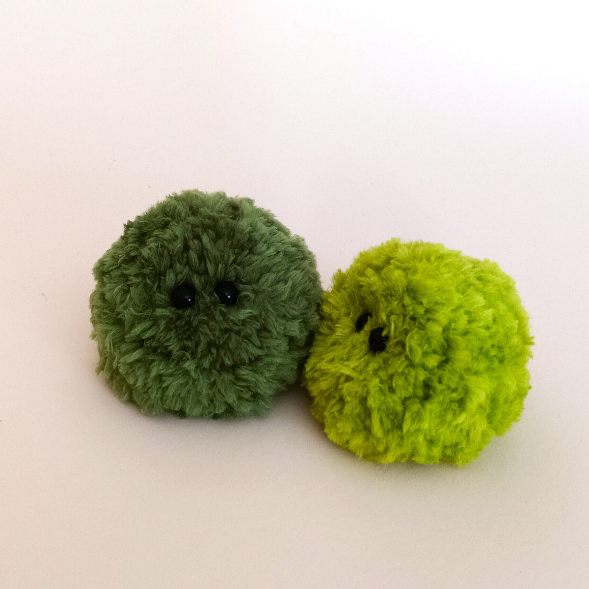 An olive green and a bright lime green fuzzy moss ball desk pet sitting closely together. Cute artificial moss pack alternative and amigurumi plush decor.
