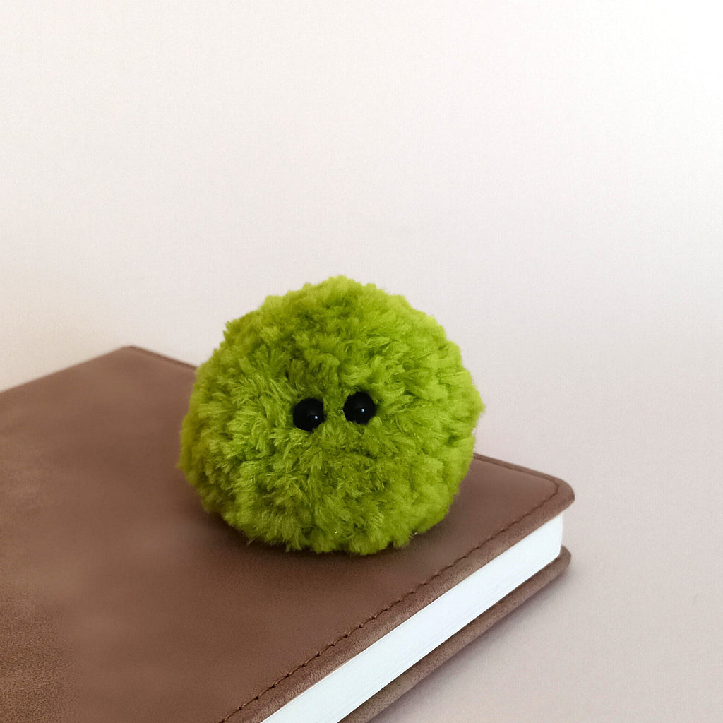 A tiny single lime green fuzzy moss ball plush sitting on a brown leather notebook. Minimalist aesthetic cottagecore desk decor and cute desk plant alternative.