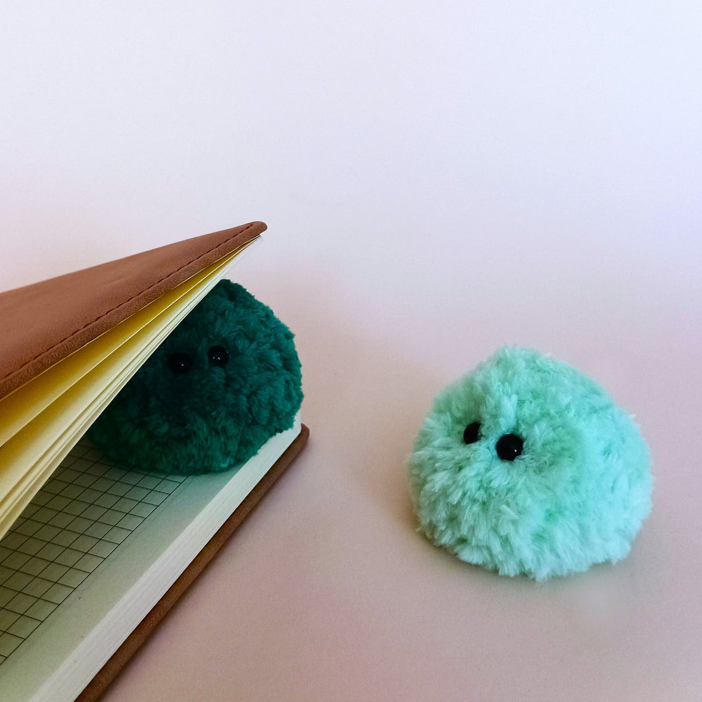 A tiny dark green fuzzy moss ball plush hiding between the pages of a brown leather planner, while a light mint green moss plush companion stands next to it. Cozy aesthetic office desk accessories and creative workspace decor.
