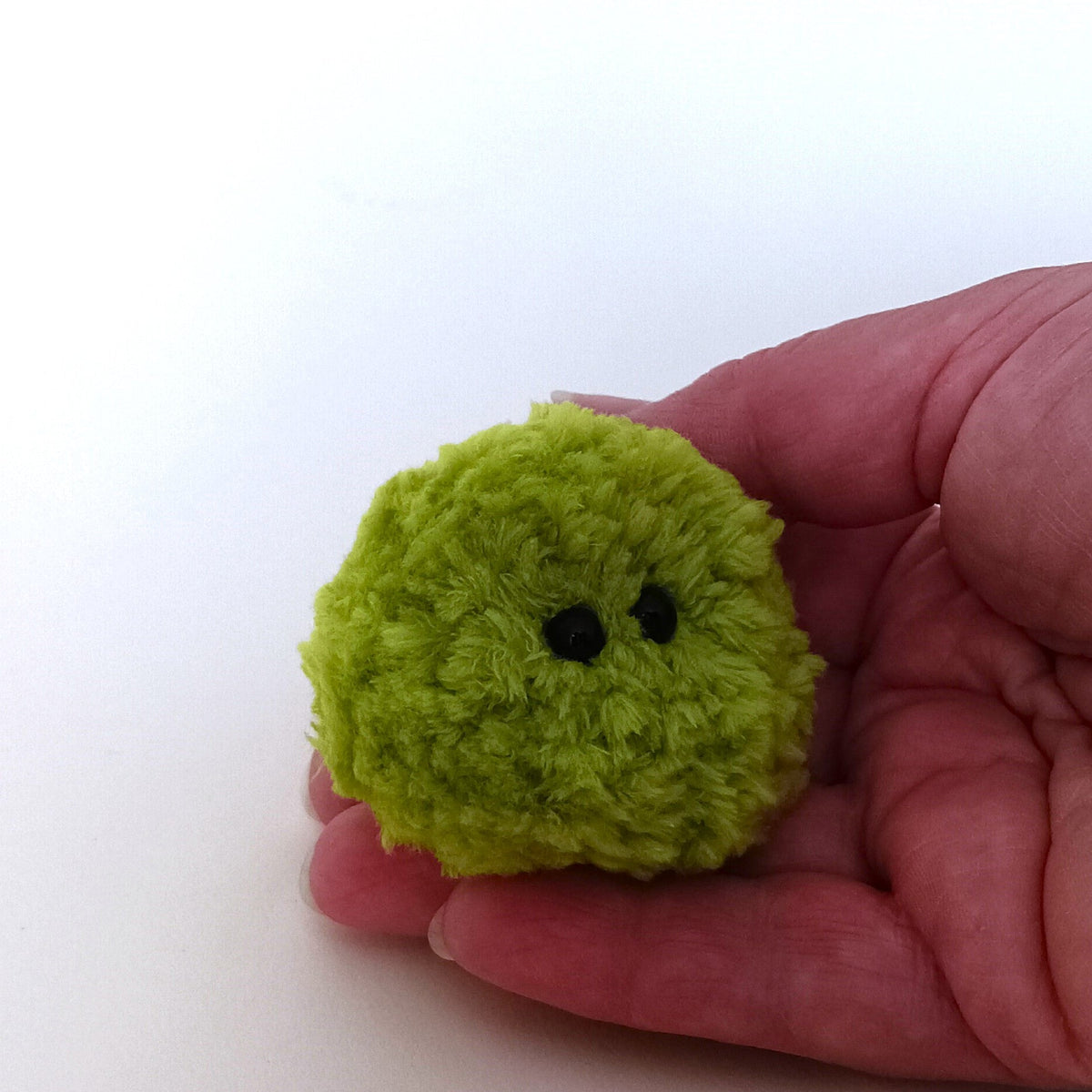 A human hand holding a tiny lime green fluffy moss ball plush, showing its miniature 2-inch pocket-sized scale. Cute handmade pocket hug gift idea.