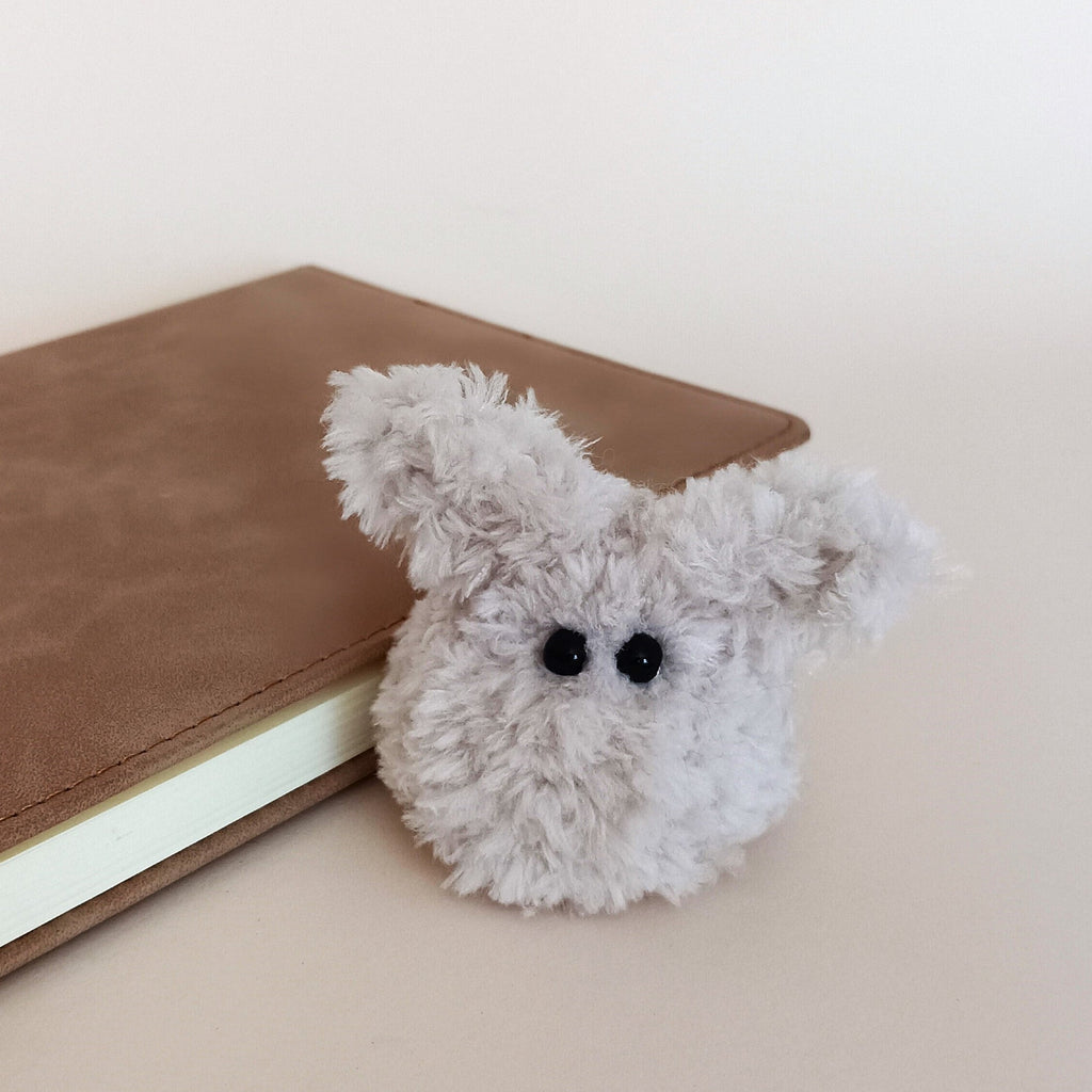A tiny handmade fuzzy bunny plush desk pet sitting next to a brown leather planner. Cozy minimalist workspace accessories and cute soft anxiety relief fidget toy for adults by PlushWhisper.
