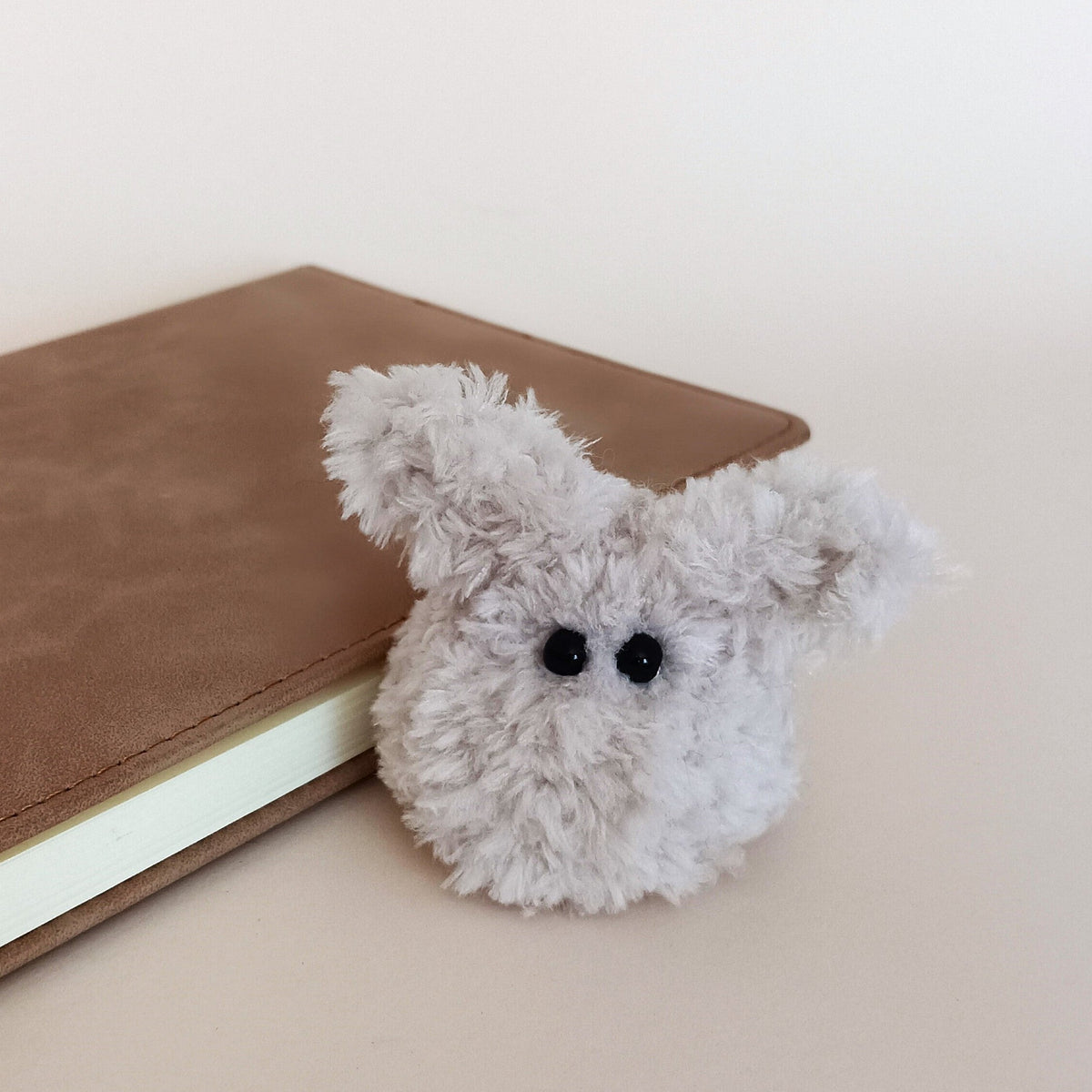 A tiny handmade fuzzy bunny plush desk pet sitting next to a brown leather planner. Cozy minimalist workspace accessories and cute soft anxiety relief fidget toy for adults by PlushWhisper.
