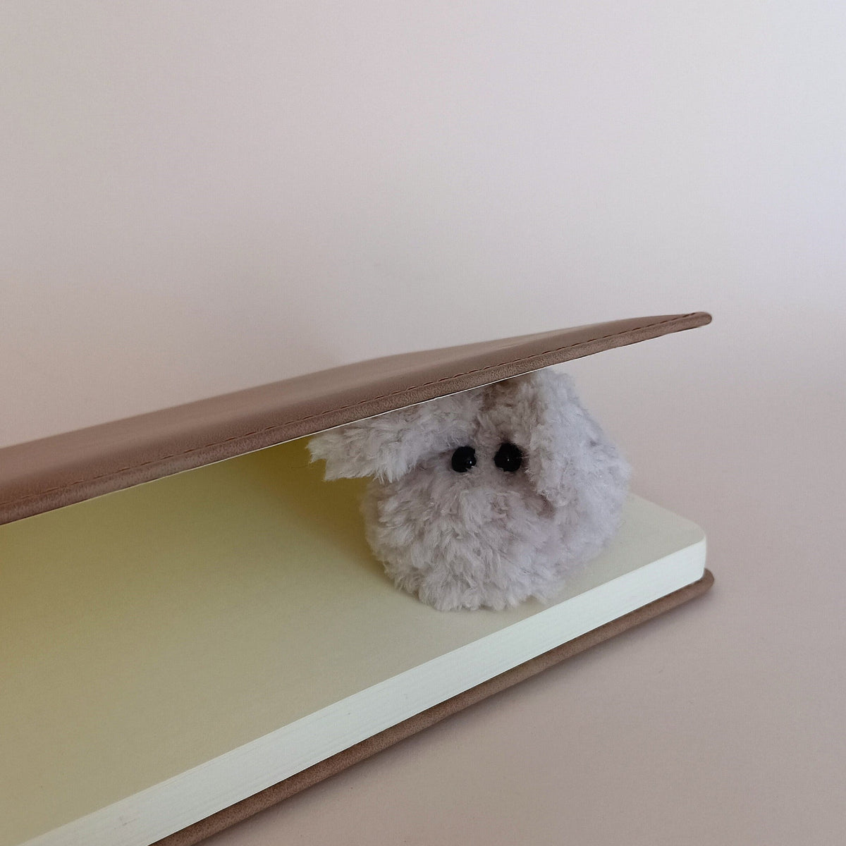 A miniature fluffy grey worry bunny pet peeking out from a brown paper notebook. Cute workspace aesthetic element and whimsical school or office stationery setup accessory.