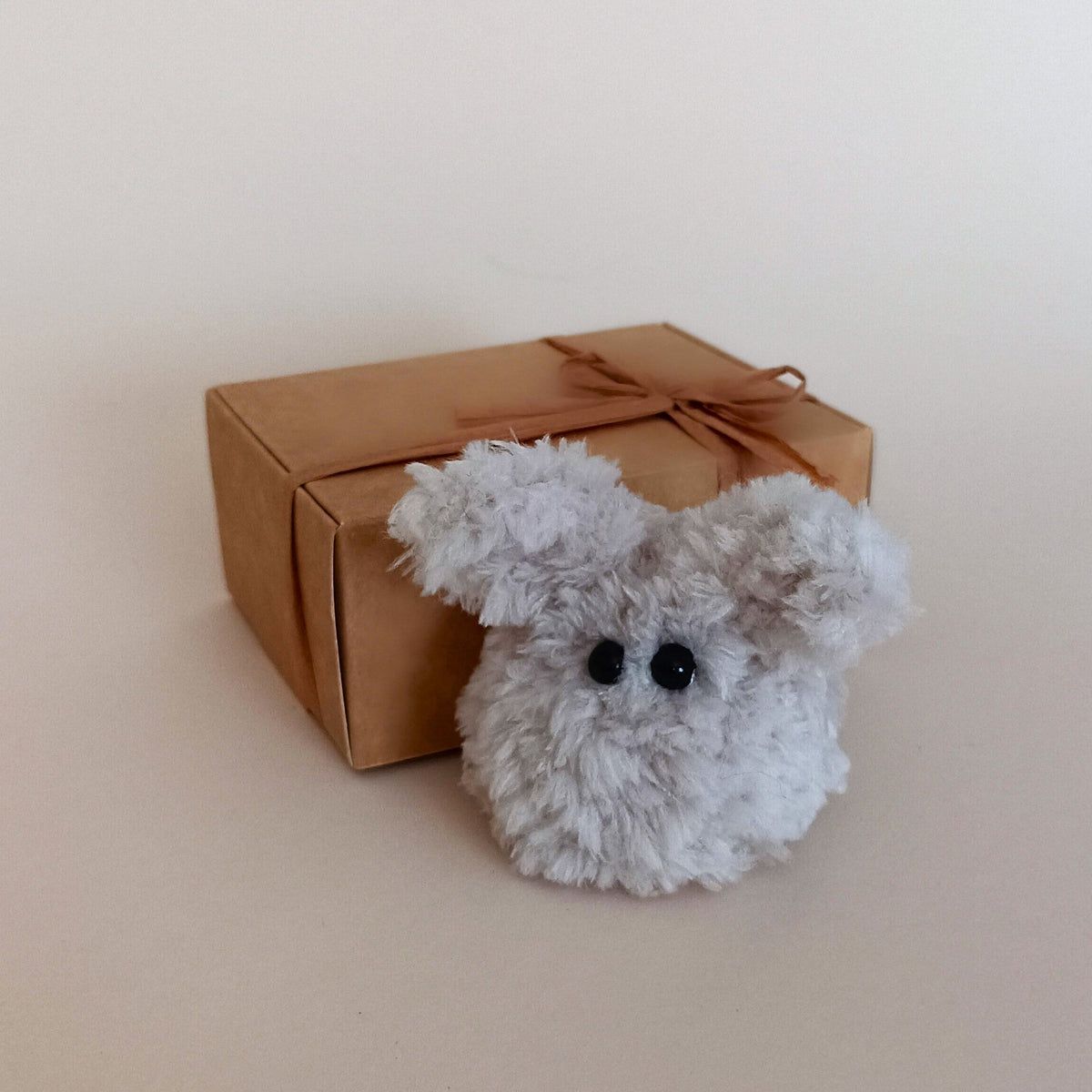A miniature handmade fuzzy grey bunny desk pet standing next to an aesthetic kraft paper gift box tied with a ribbon bow. Fluffy pocket size worry pet packaging display and unique small holiday gift box idea for coworkers by PlushWhisper.