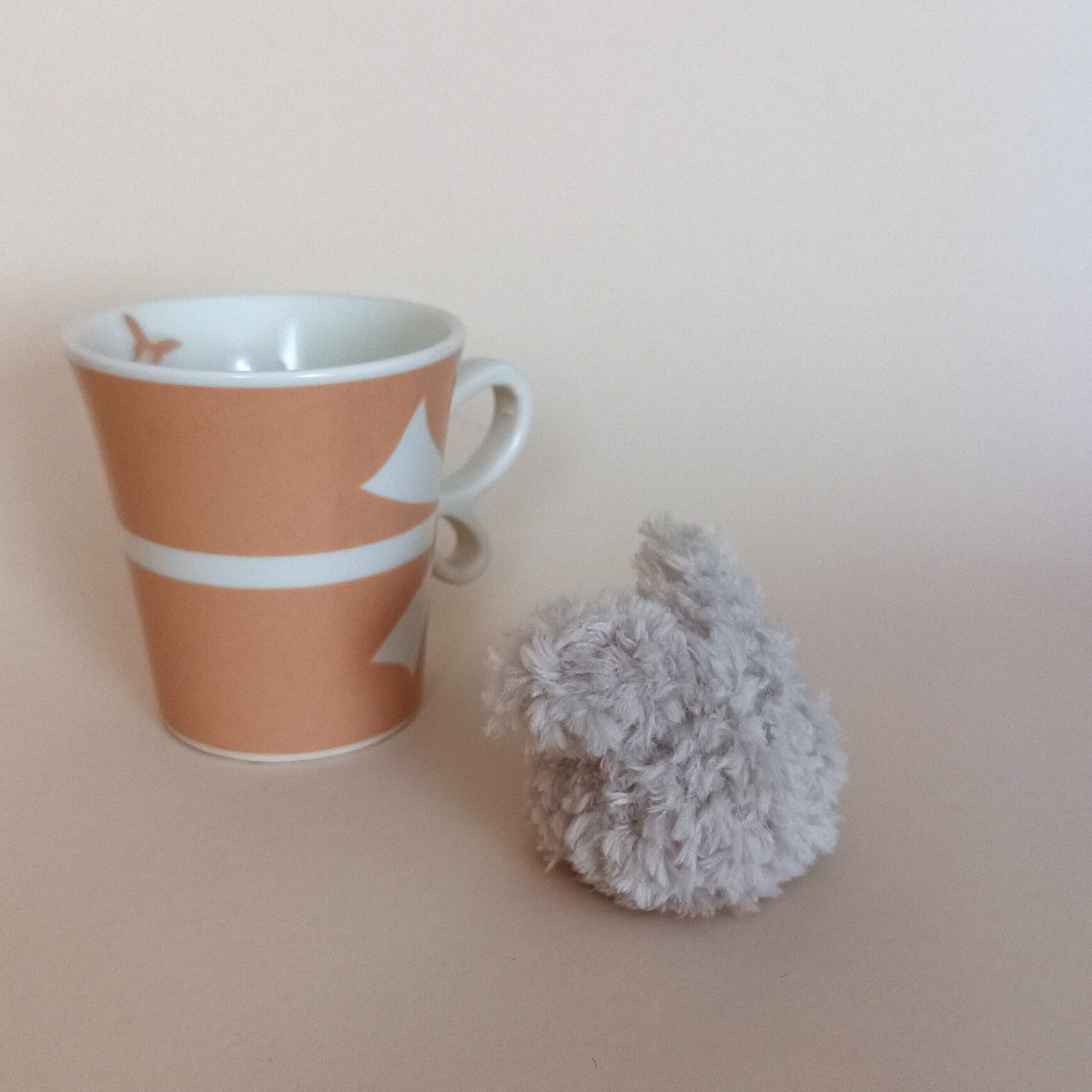 Miniature grey fuzzy bunny desk pet sitting on a table next to cozy ceramic coffee cups. Aesthetic hygge kitchen decor accent and unique small gift box idea for coffee lovers.
