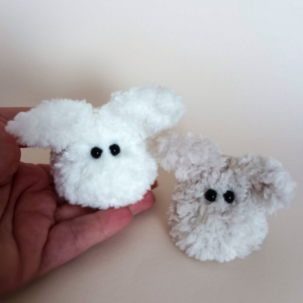 Two miniature ultra fluffy handmade worry pet bunnies in white and light grey colors sitting on a person's palm to show the pocket size scale. Small cute autumn or winter gift idea by PlushWhisper.