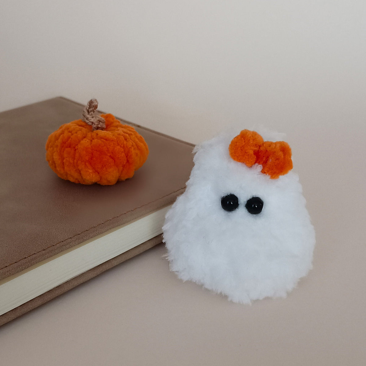 A small white fluffy ghost-shaped ornament or plush with black bead eyes and an orange bow, placed beside a brown book and a small orange pumpkin on a beige surface.