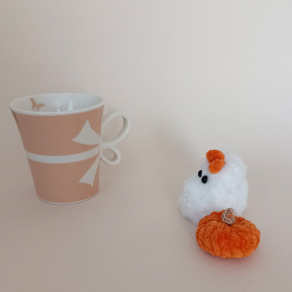 Tiny white crochet ghost with orange bow and handmade orange pumpkin beside a coffee cup, cozy Halloween desk decor