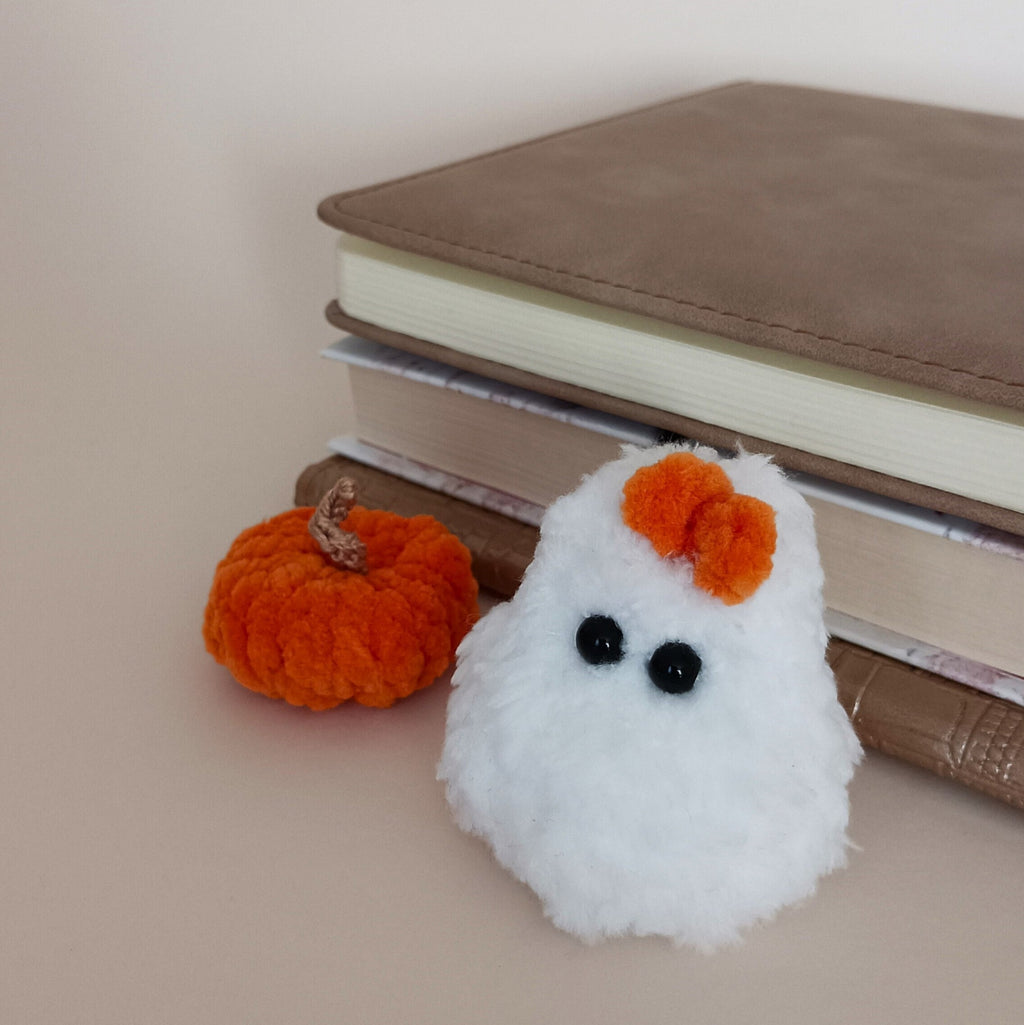 Tiny white crochet ghost with orange bow and matching orange pumpkin displayed beside a stack of books, cozy Halloween shelf decor