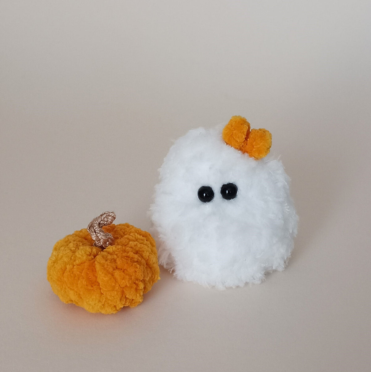 Tiny white crochet plush ghost with a small orange bow sitting beside a handmade yellow-orange pumpkin, cute aesthetic Halloween desk decor and emotional support companion by PlushWhisper.