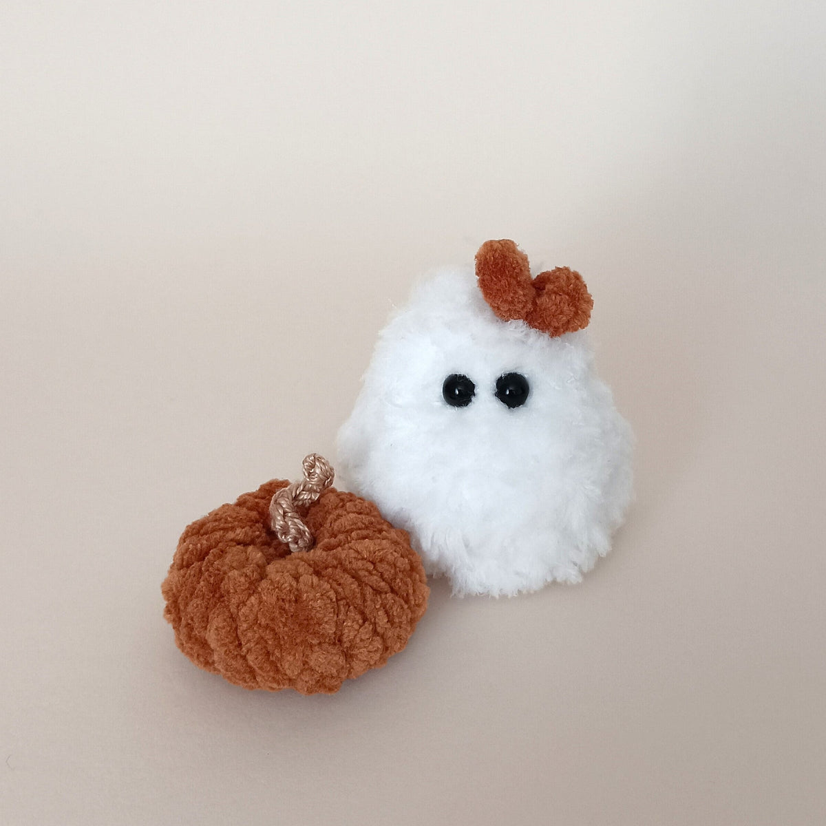 Small white handmade plush ghost with a brown bow beside a terracotta brown crochet pumpkin, cozy fall workspace aesthetic and cute pocket-sized desk pet by PlushWhisper.