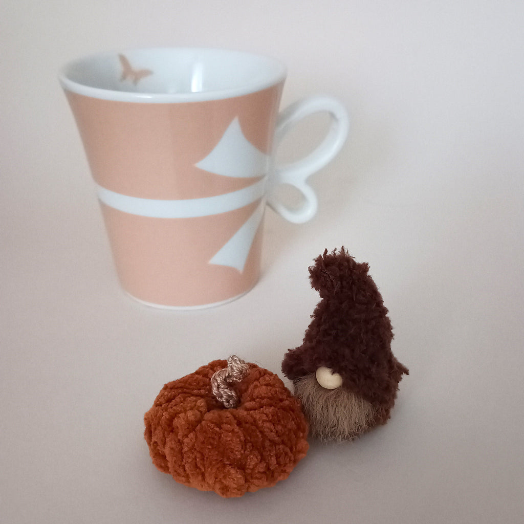 A tiny handmade coffee gnome plush sitting next to a miniature orange crocheted pumpkin and a pastel pink mug on a light background. Cozy autumn aesthetic home decor and small fall themed gift ornament for coffee lovers.