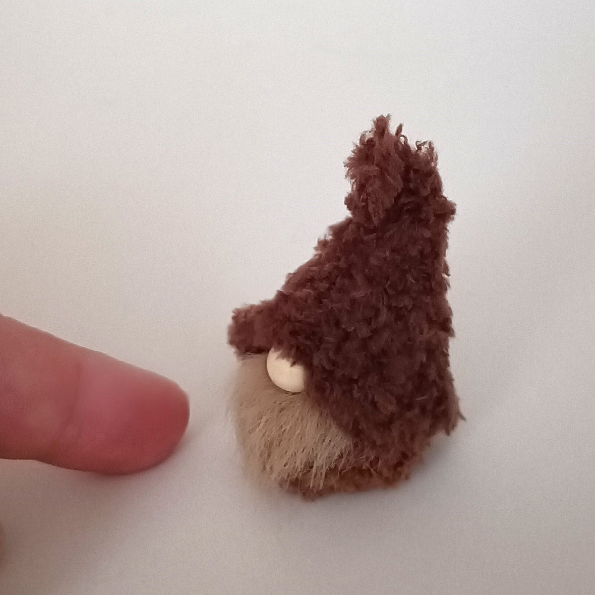 A finger pointing at a tiny handmade espresso brown coffee gnome plush to demonstrate its miniature pocket size. Cute primitive desk pet and small minimalist office companion for coffee lovers.