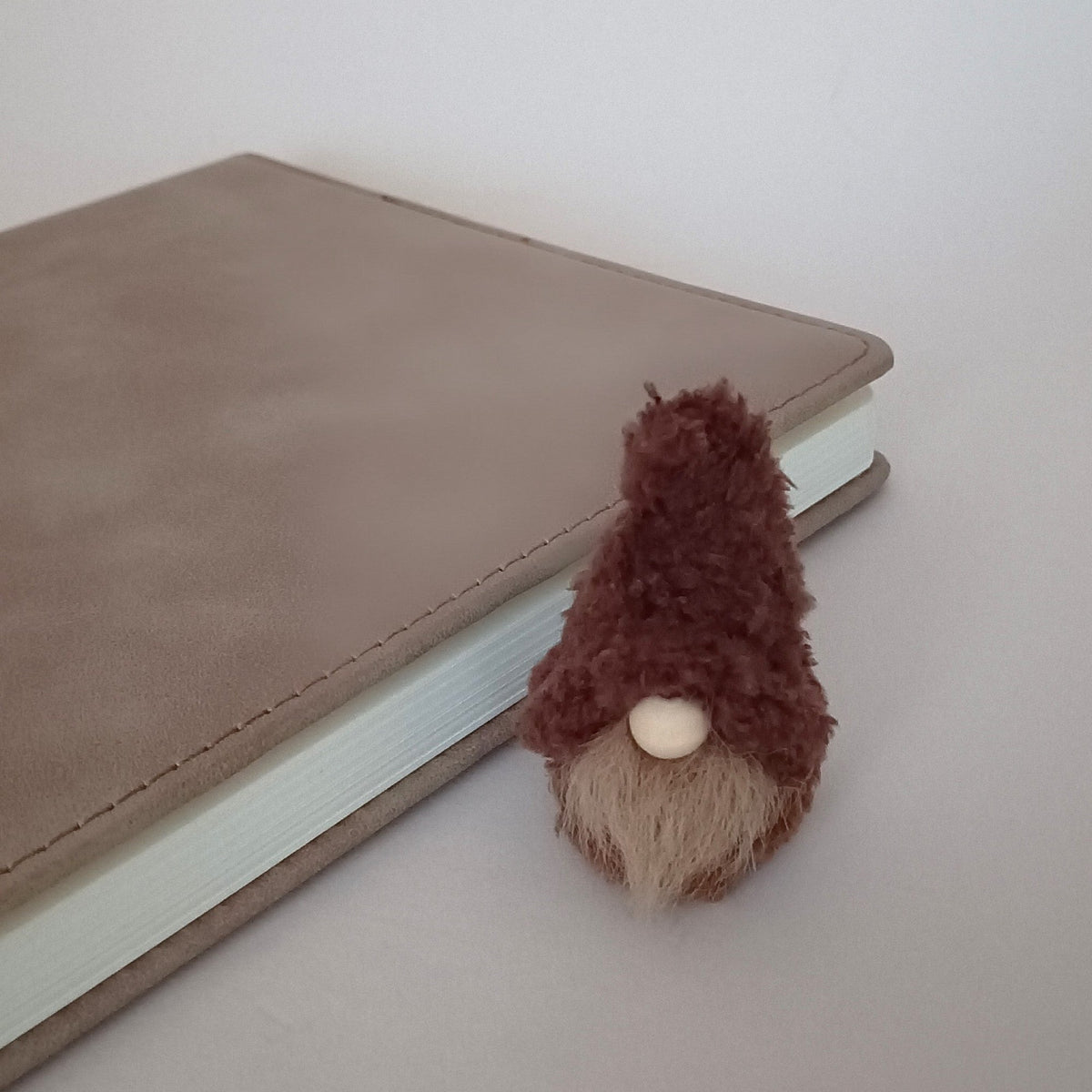 A miniature handmade coffee gnome plush with a fuzzy brown hat sitting next to a gray leather planner on a desk. Cute office workspace companion and tiny primitive pocket buddy gift for coffee lovers.
