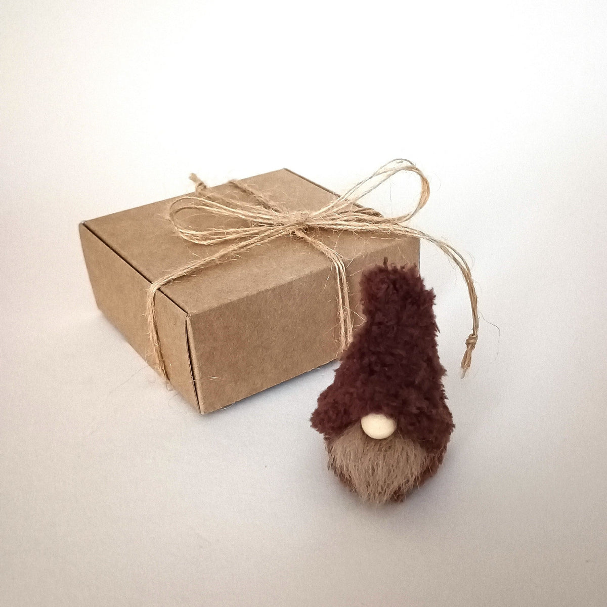 A tiny handmade rich espresso dark brown coffee gnome plush standing next to a kraft paper gift box tied with a twine bow. Minimalist aesthetic desk pet ornament and small coworker birthday gift for coffee lovers.