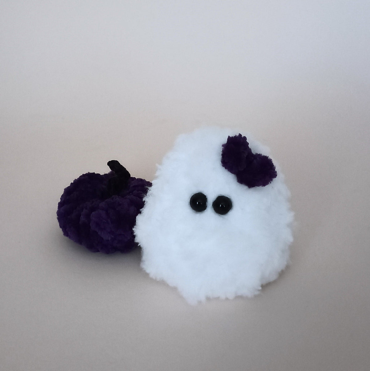 White handmade ghost with a purple bow beside a matching purple pumpkin, a tiny Halloween decor set made from soft fluffy yarn
