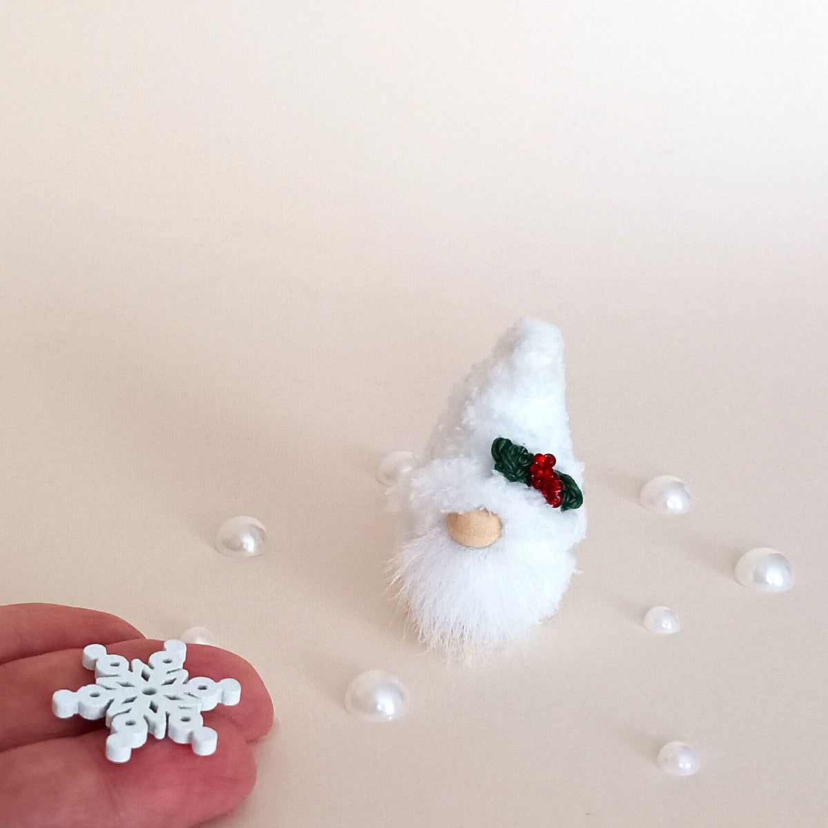 Miniature 2-inch Christmas pocket gnome next to pearl beads and a hand holding a tiny wooden snowflake, creating a magical winter aesthetic.