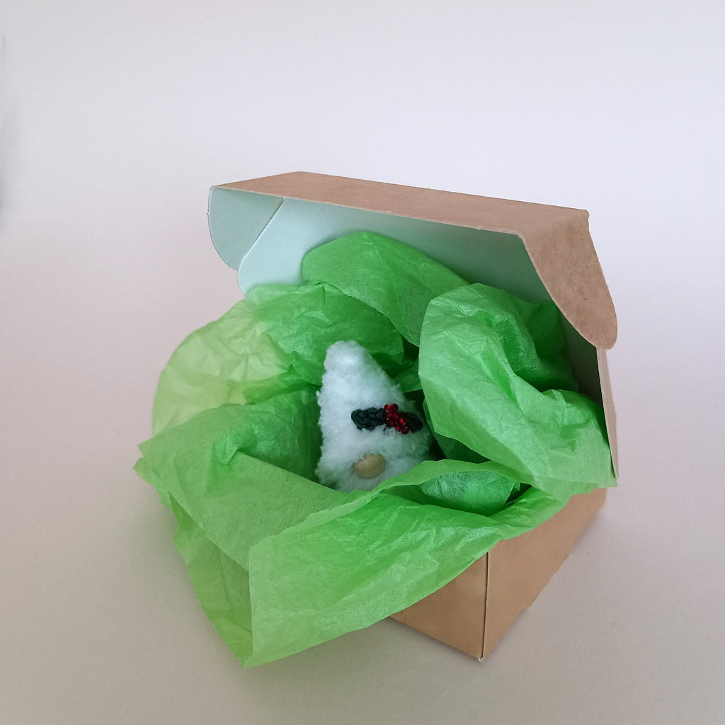 Small white plush Christmas gnome inside an open craft gift box with festive green tissue paper, showing eco-friendly holiday unboxing experience.