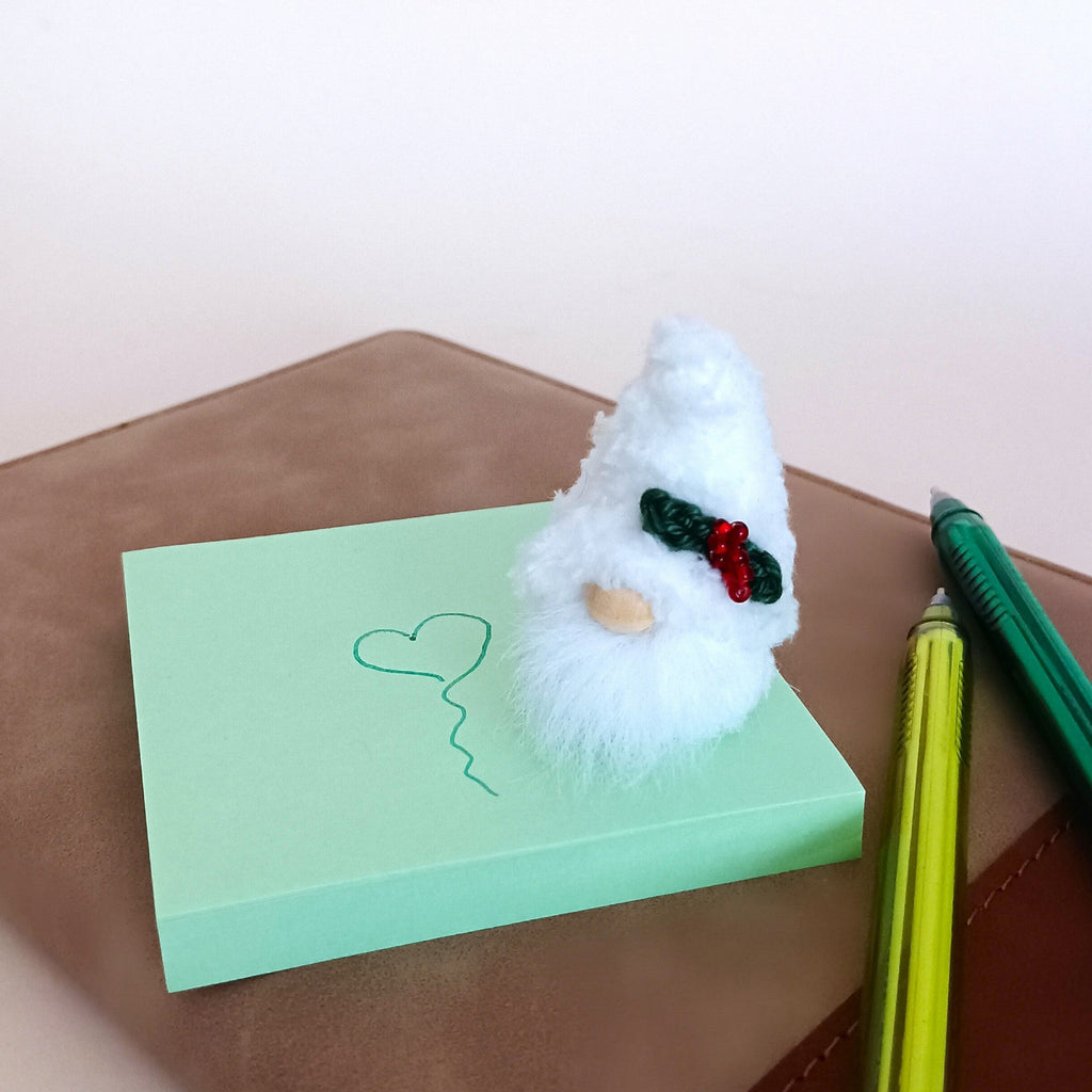 Small handmade Christmas gnome ornament standing on a green sticky note with a drawn heart, showing cute tabletop office stationery decoration.