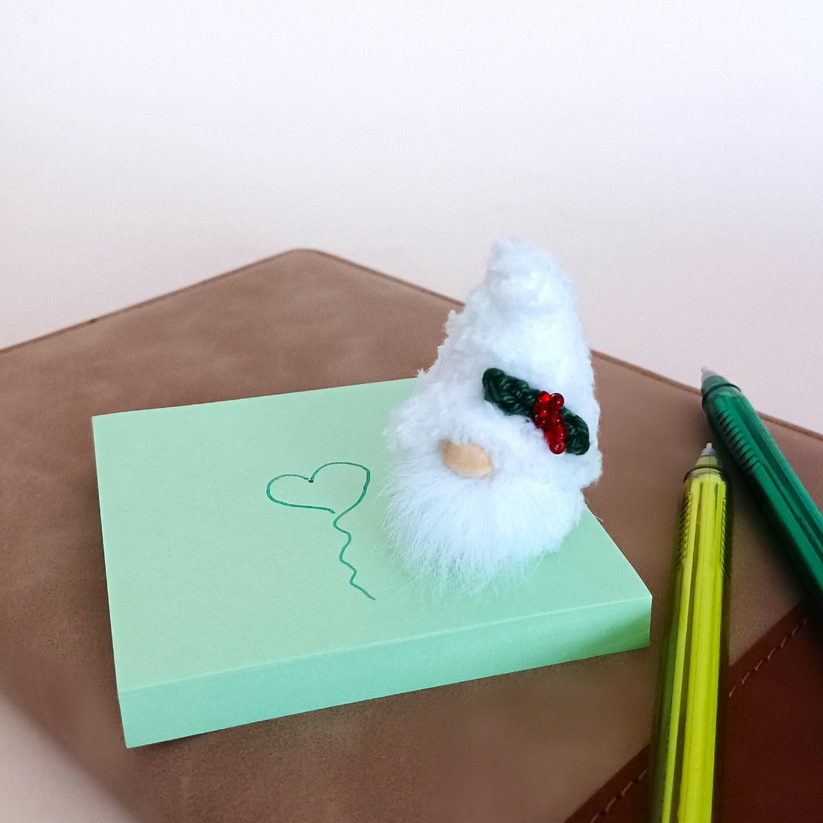 Small handmade Christmas gnome ornament standing on a green sticky note with a drawn heart, showing cute tabletop office stationery decoration.