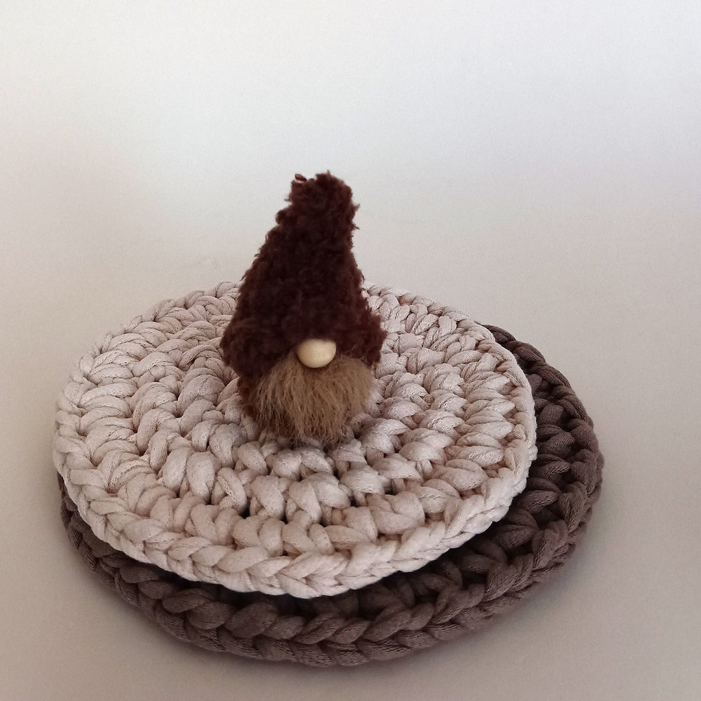 A miniature handmade espresso brown coffee gnome plush standing on stacked crocheted round coasters. Cozy minimalist home coffee bar decor and a tiny aesthetic shelf ornament gift for coffee lovers.