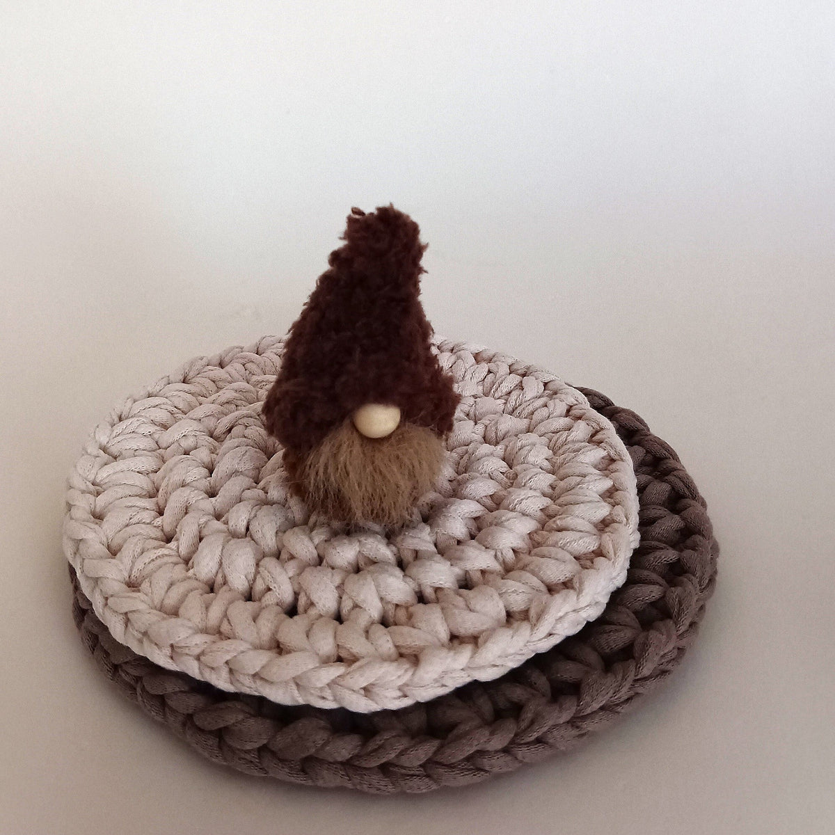 A miniature handmade espresso brown coffee gnome plush standing on stacked crocheted round coasters. Cozy minimalist home coffee bar decor and a tiny aesthetic shelf ornament gift for coffee lovers.