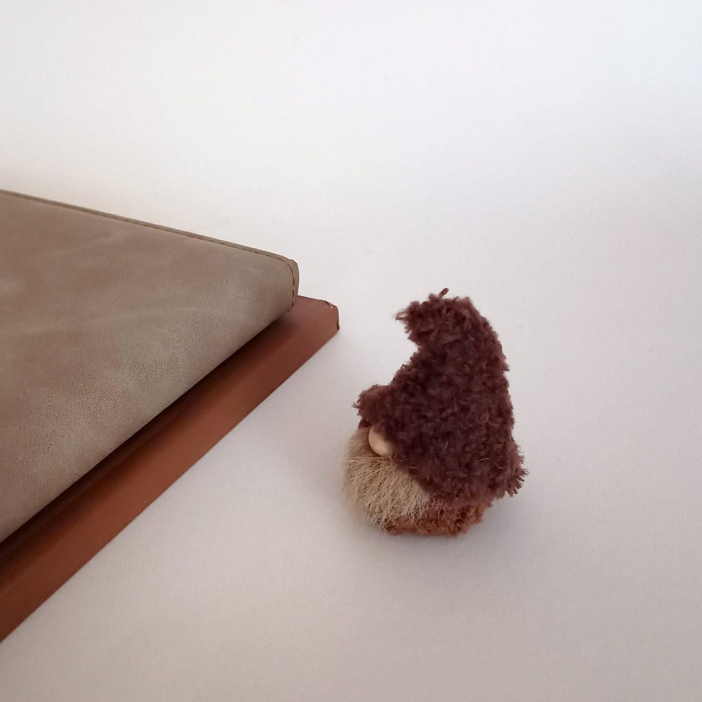 A miniature handmade coffee gnome plush seen from the side, sitting next to a gray leather planner on a neutral desk. Cute office workspace companion and tiny primitive pocket buddy gift for coffee lovers.