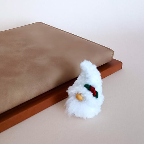 Christmas pocket gnome plush with a fluffy white beard and holly leaf hat, sitting next to a beige leather notebook as cozy winter office desk decor.
