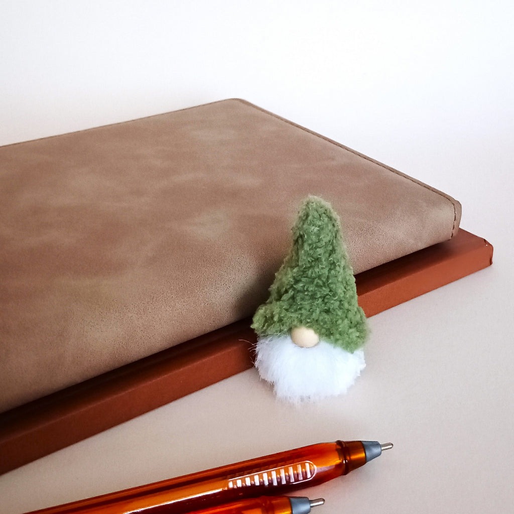 Miniature green Christmas forest gnome plush with a fluffy white beard, sitting next to a brown leather notebook and pens as cozy winter office desk decor.