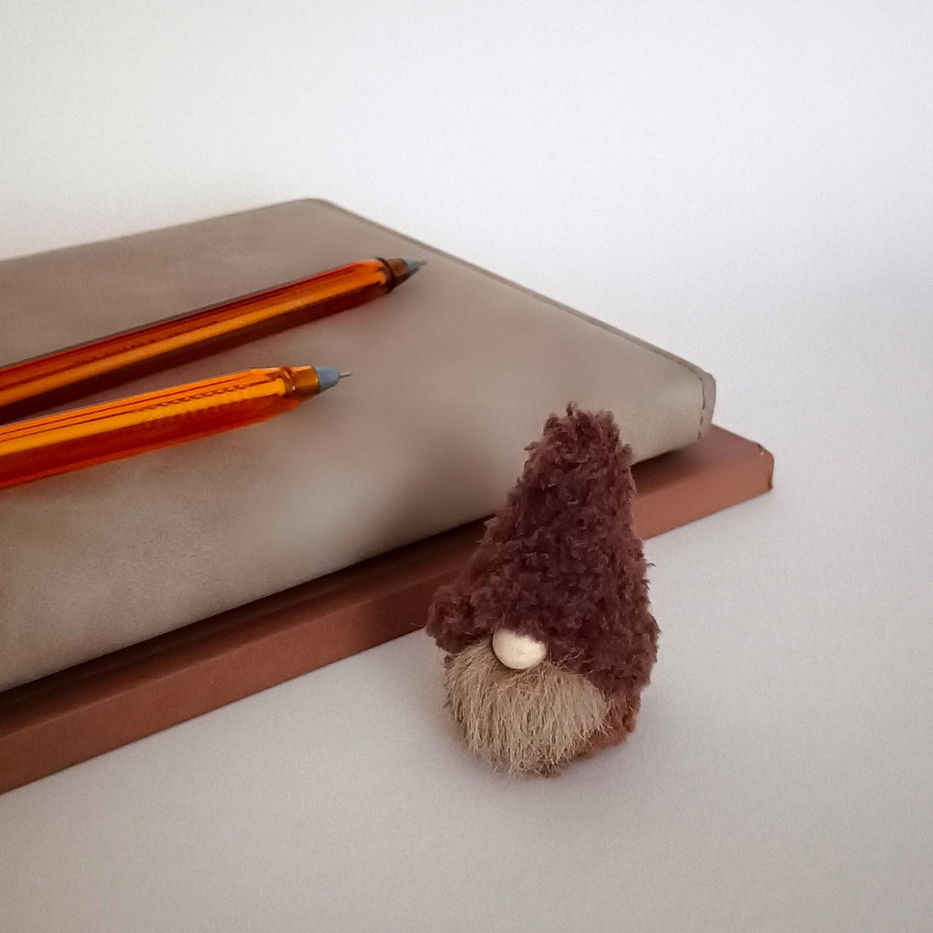 A small handmade cappuccino brown coffee gnome plush sitting next to a gray leather-bound planner and two amber ballpoint pens. Cute office workspace companion and tiny aesthetic desk pet gift for coffee lovers.