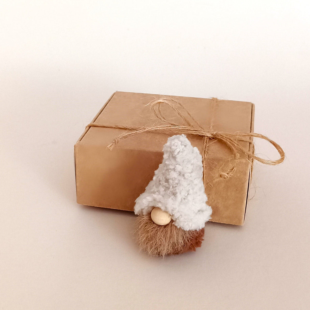 A tiny handmade cream color coffee gnome plush sitting in front of a kraft paper gift box tied with a twine bow. Aesthetic packaging for a small coworker gift and miniature pocket companion for coffee lovers.
