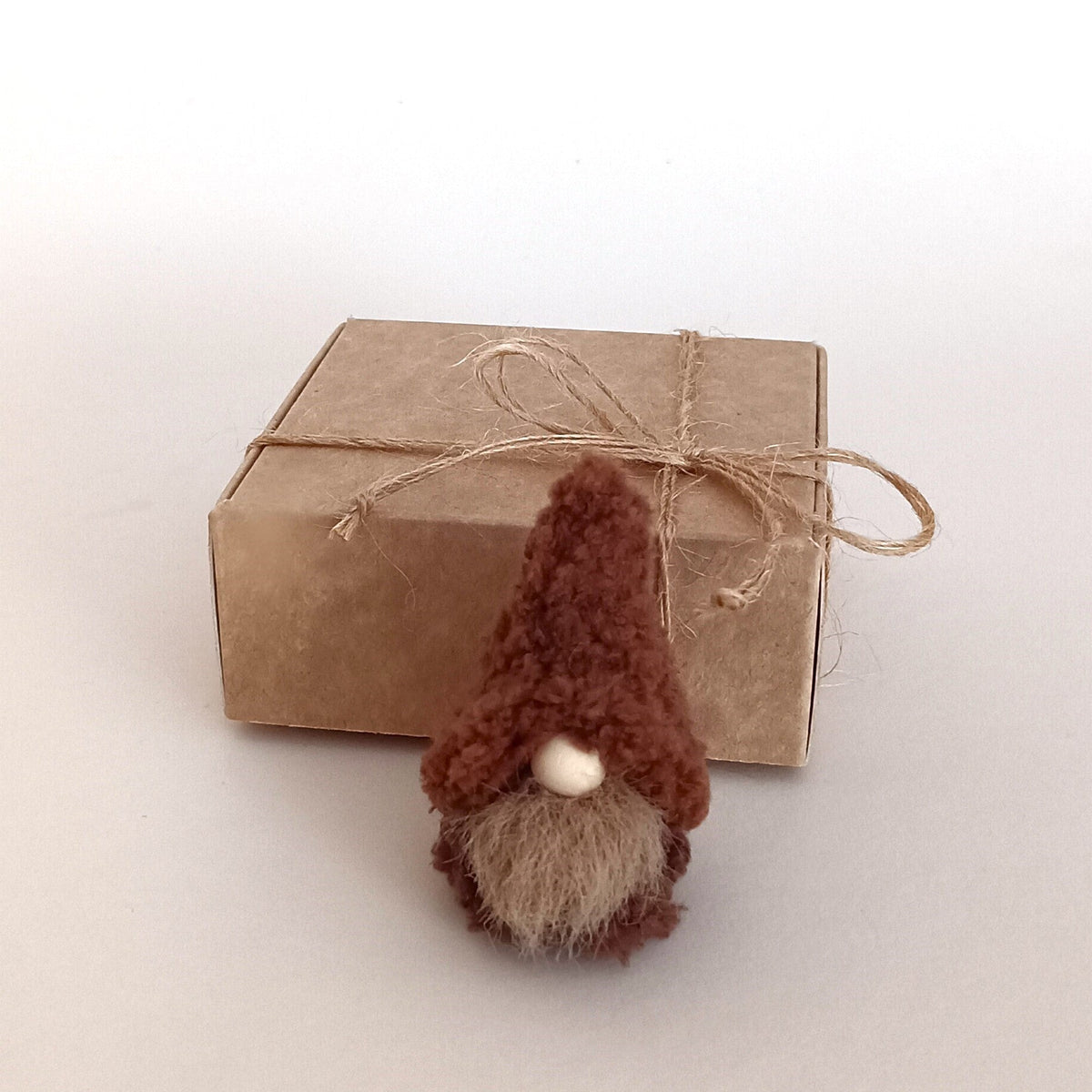 A small handmade cappuccino brown coffee gnome plush standing in front of a kraft paper gift box tied with a twine ribbon. Cozy primitive desk pet and miniature secret santa holiday gift idea for coffee lovers.
