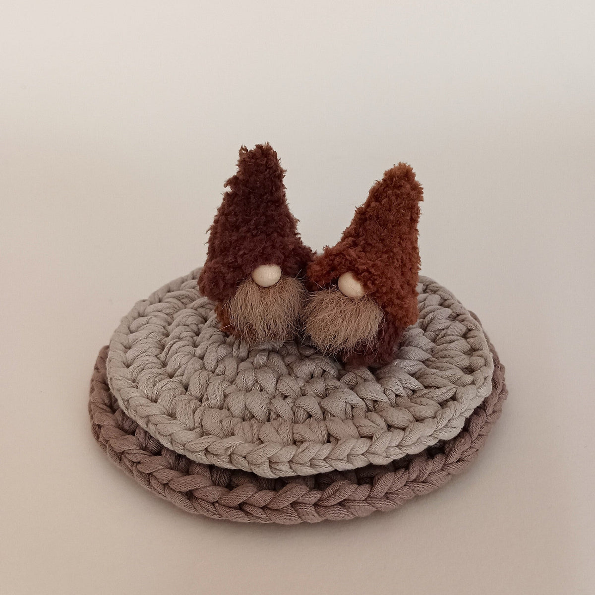 Two small handmade coffee gnome plushies in cappuccino and espresso brown colors sitting on stacked crocheted coasters. Cozy miniature coffee bar decor and modern kitchen shelf ornaments for coffee lovers.