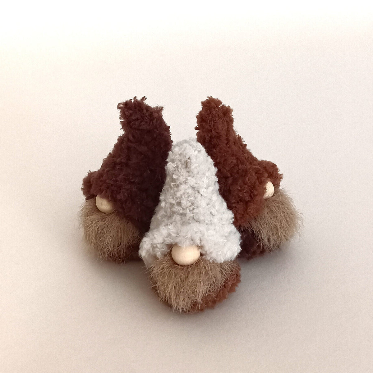 A close-up of three miniature handmade coffee gnome plushies in cream, cappuccino, and espresso brown colors grouped together on a white background. Cute set of tiny minimalist desk buddies and small holiday gifts for coffee lovers.