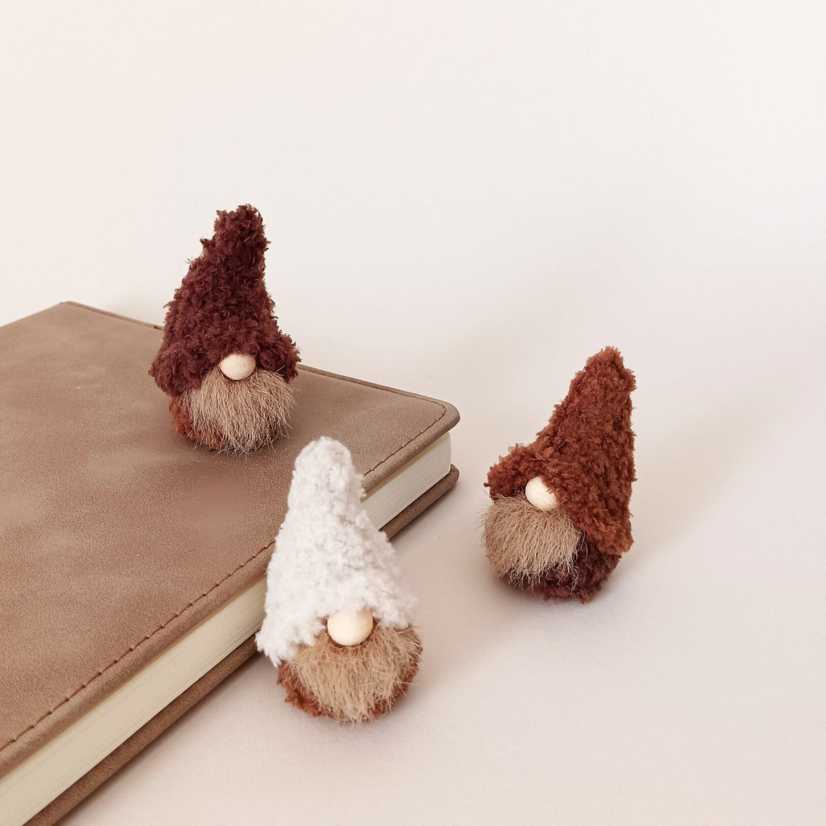 Three tiny handmade coffee gnome plushies in cream, cappuccino, and espresso brown colors sitting on and next to a brown leather-bound book. Cozy minimalist workspace decor and miniature desk buddy gifts for adults.