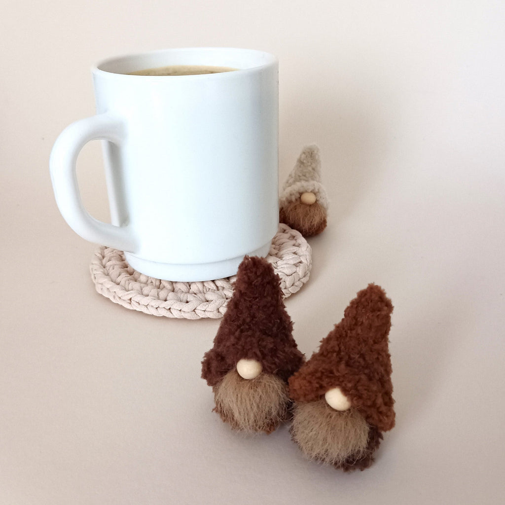 Three tiny handmade coffee gnome plushies in cream, cappuccino, and espresso brown colors standing around a white coffee mug on a neutral background. Cozy minimalist home office decor and aesthetic kitchen coffee station accessories for adults.